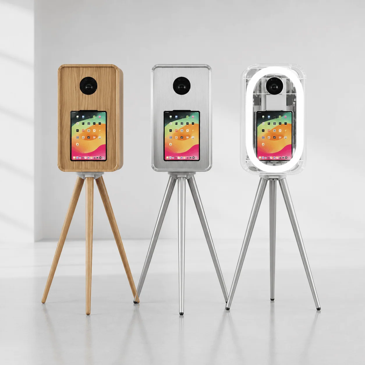Three iPad photo booth shells side by side: walnut wood enclosure, brushed aluminum enclosure, and clear acrylic enclosure with integrated LED ring light